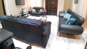 Una sala de estar con dos sofás negros, un televisor de pantalla plana, una mesa de centro y una puerta con pomo decorativo.