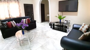 Una sala de estar con un sofá negro, una mesa de centro de vidrio y un televisor montado en la pared.