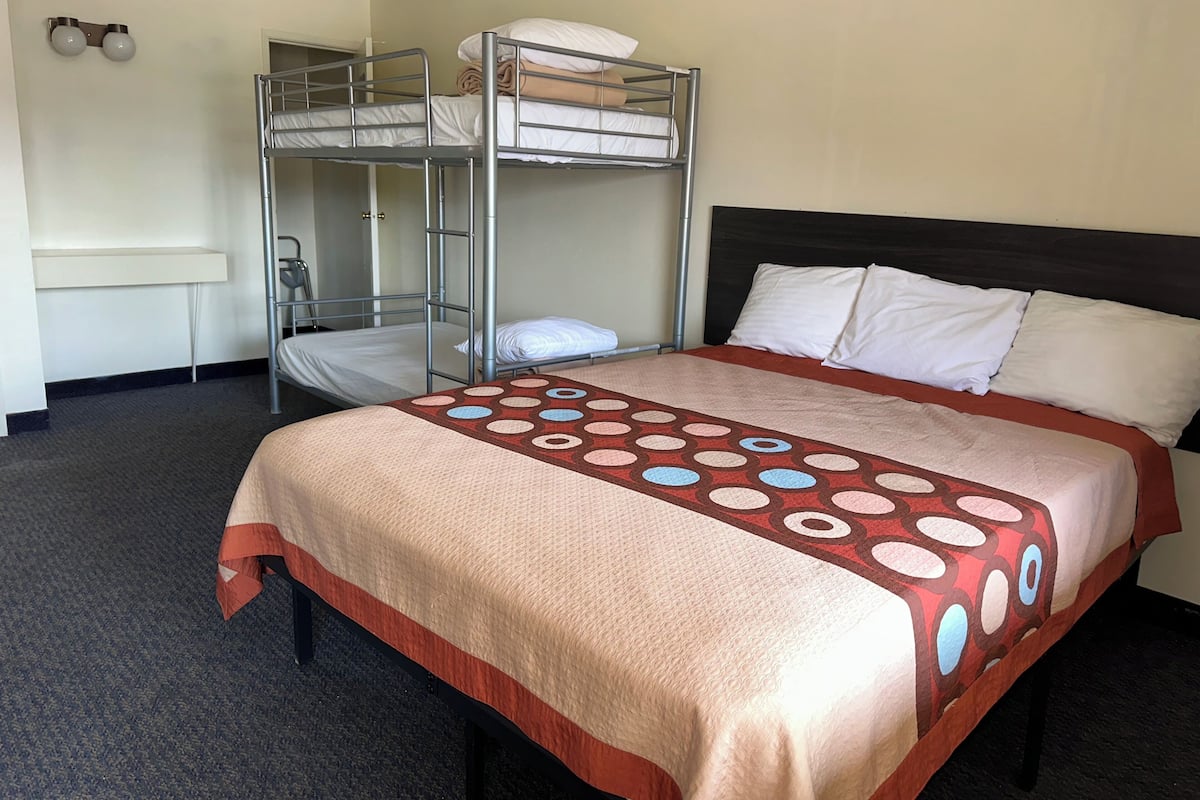 A hotel room with a bunk bed, a single bed, a patterned bedspread, and a wall-mounted light fixture.