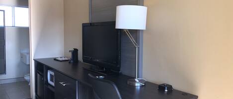 Habitación de hotel compacta con escritorio de madera oscura, televisor de pantalla plana, lámpara moderna, una silla y un pequeño cesto de basura.