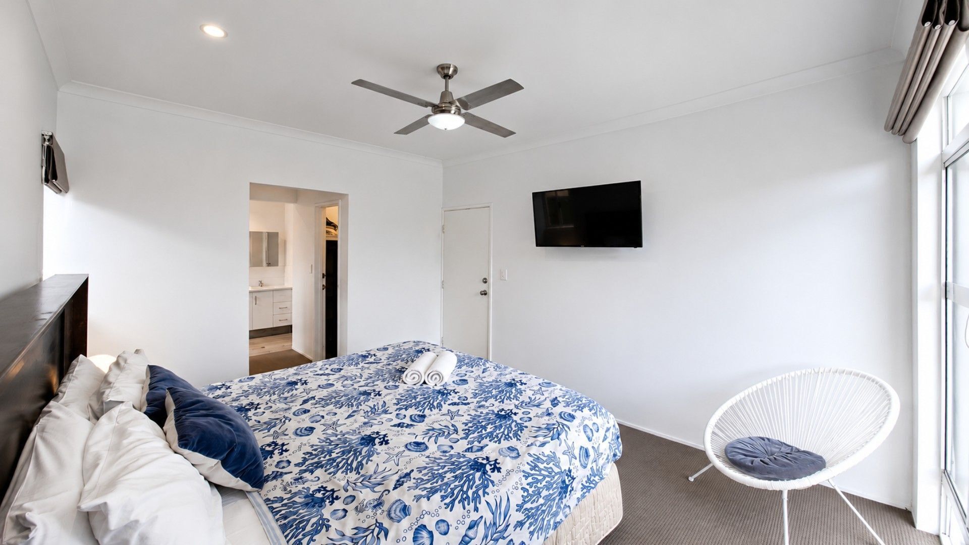 Une chambre à coucher comprenant un lit, une chaise blanche, un ventilateur de plafond, un téléviseur et une porte donnant sur une salle de bain.