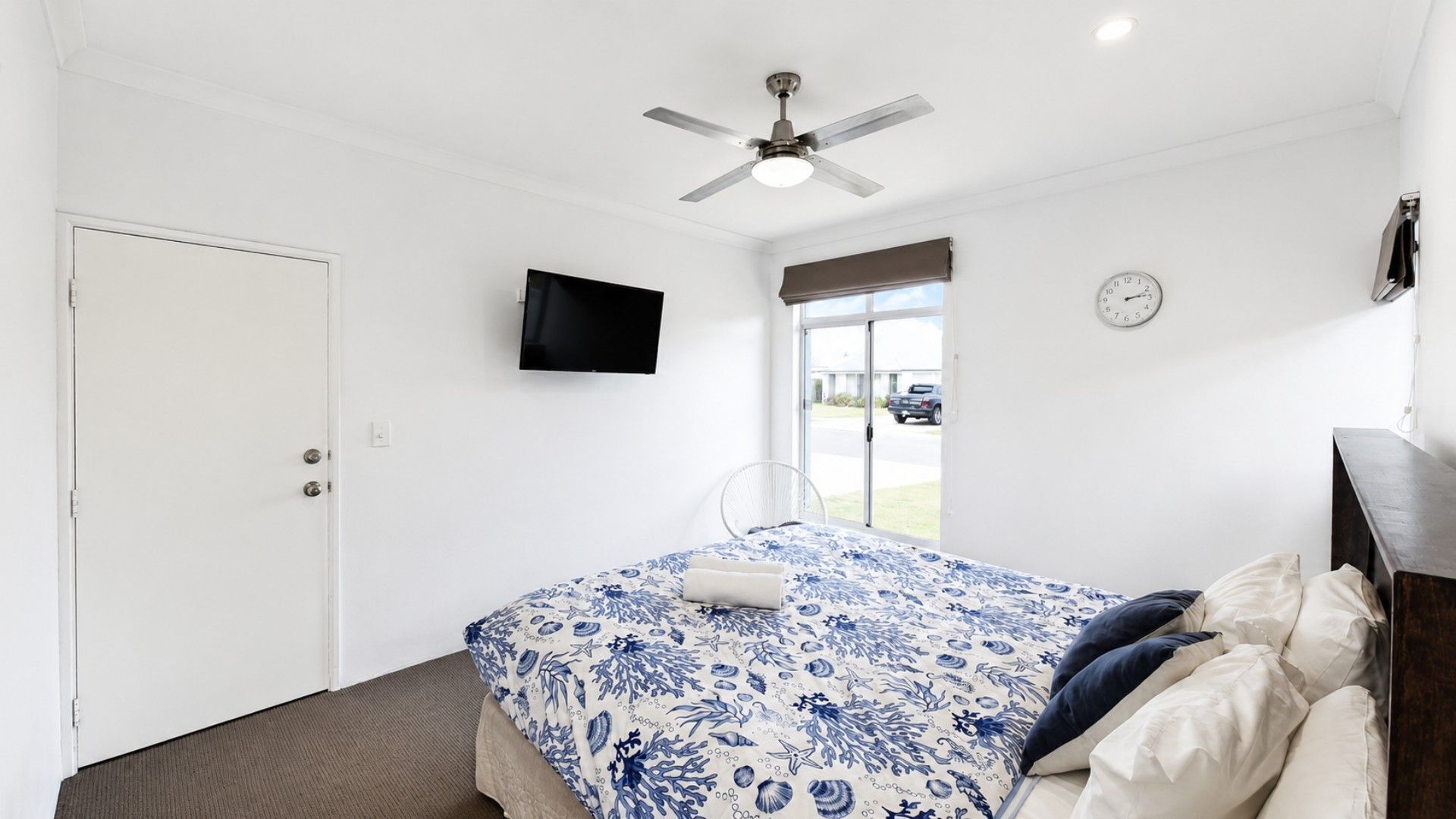A bedroom with a bed, a ceiling fan, a TV, a clock, and a window.
