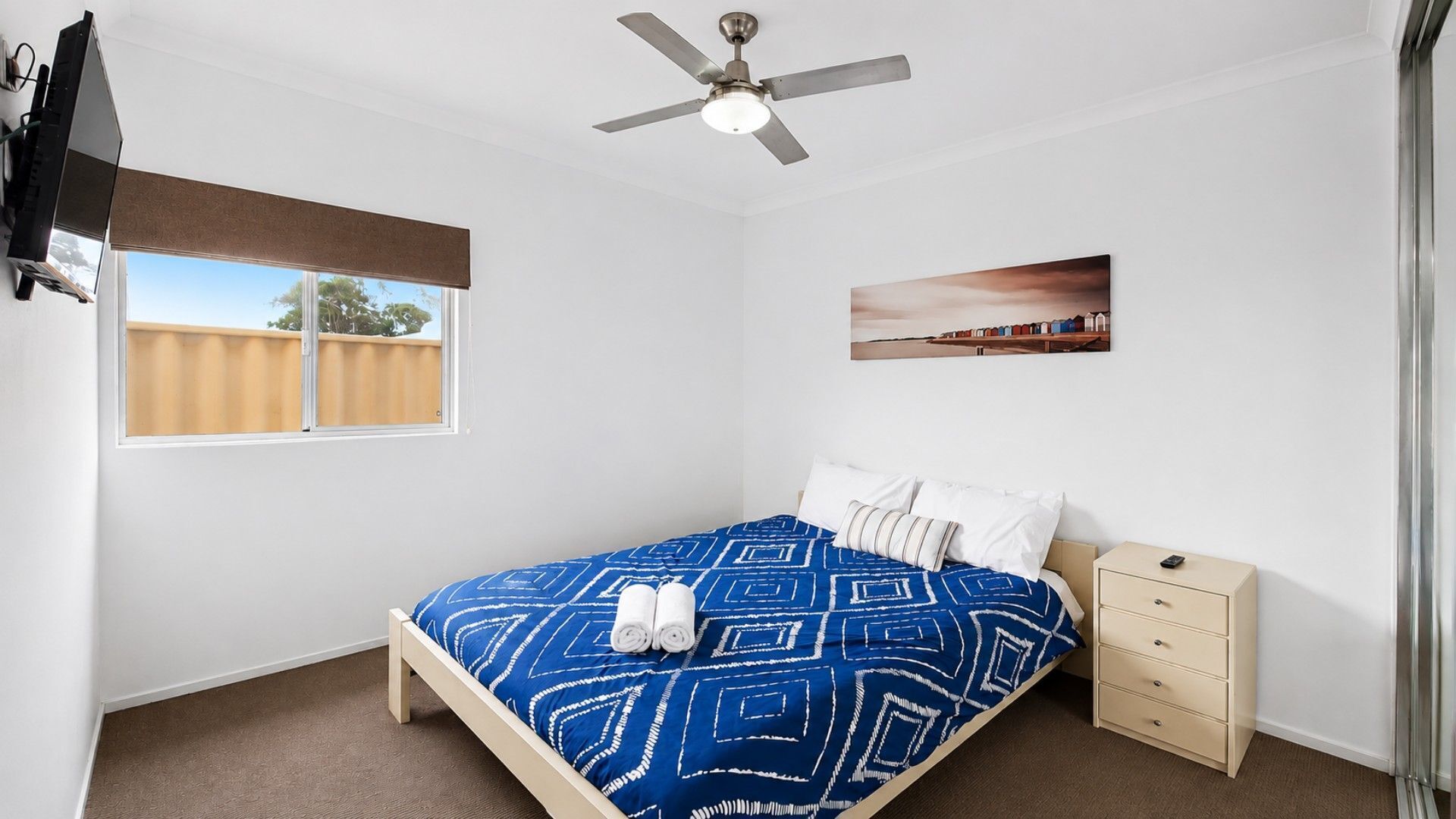 A bedroom with a bed, a nightstand, a ceiling fan, a window with blinds, and a wall-mounted picture.