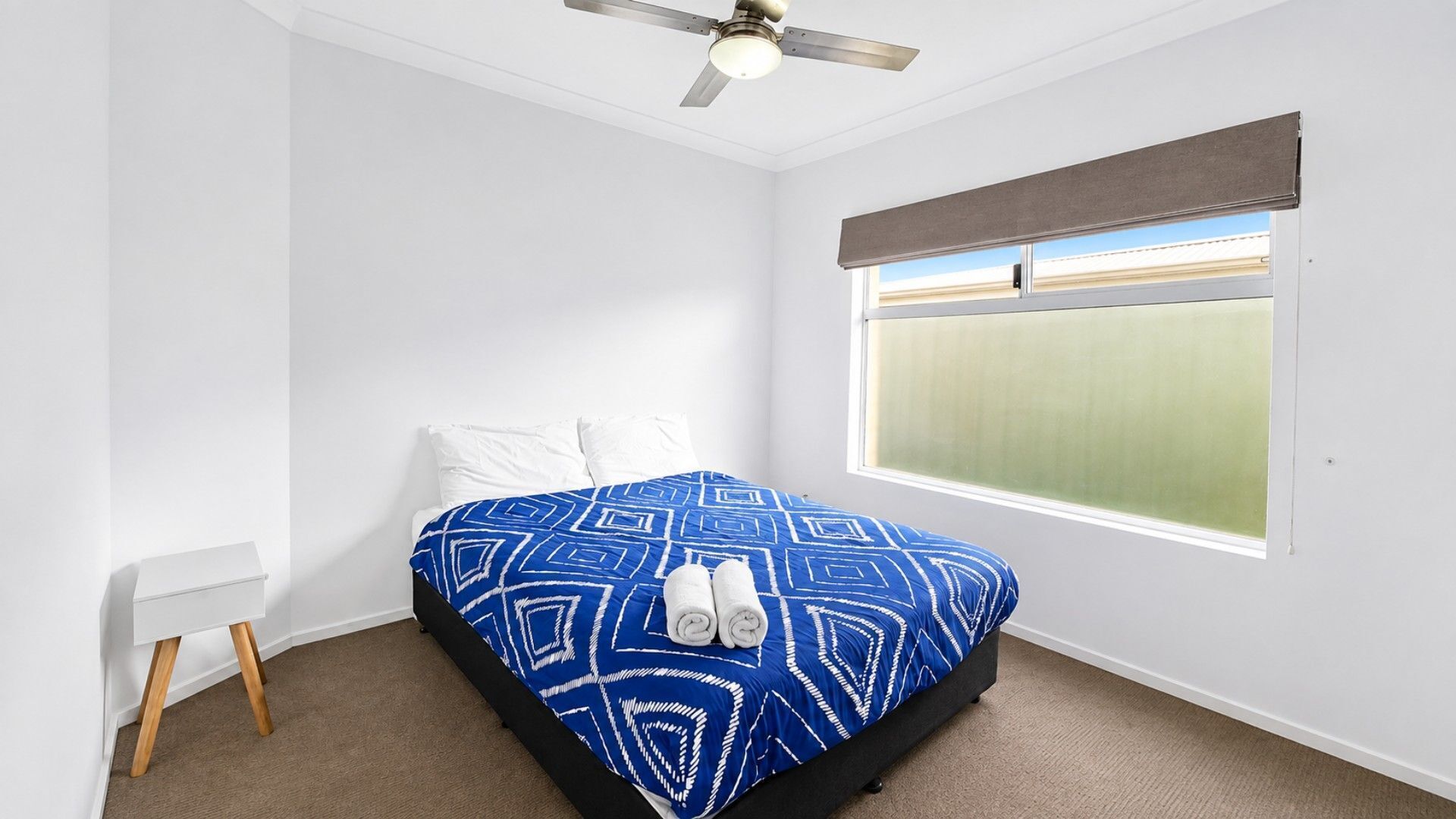 A bedroom with a bed, a ceiling fan, a window with blinds, and a small bedside table.