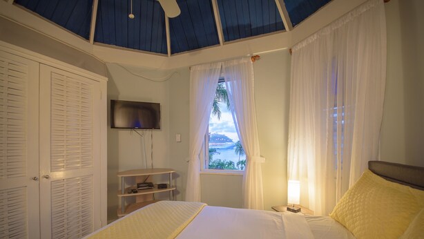 A bedroom with a bed, a TV mounted on the wall, a window with curtains, and a view of the sea.