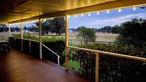 Een overdekt buitengebied met een houten terras, sfeerverlichting en uitzicht op een grasveld en bomen.
