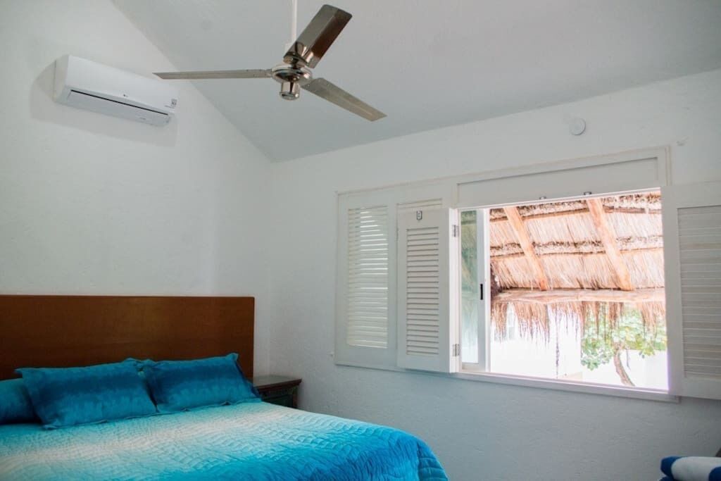 A bedroom with a bed, an air conditioning unit, a ceiling fan, and a window with shutters.