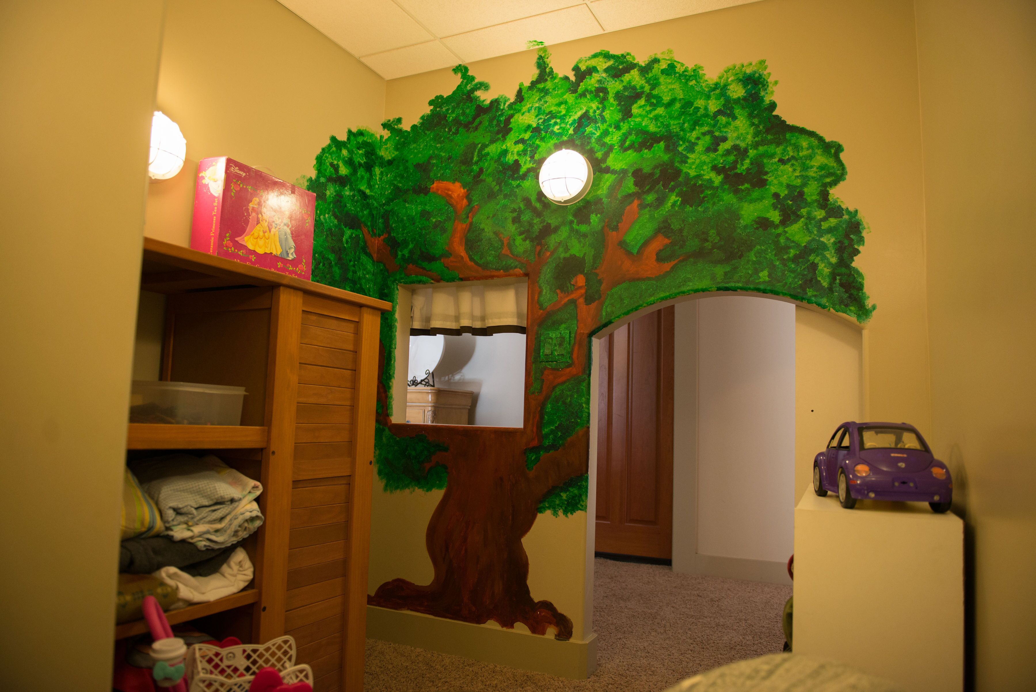 A child's playroom with a tree mural, a wooden shelf, and a toy car.