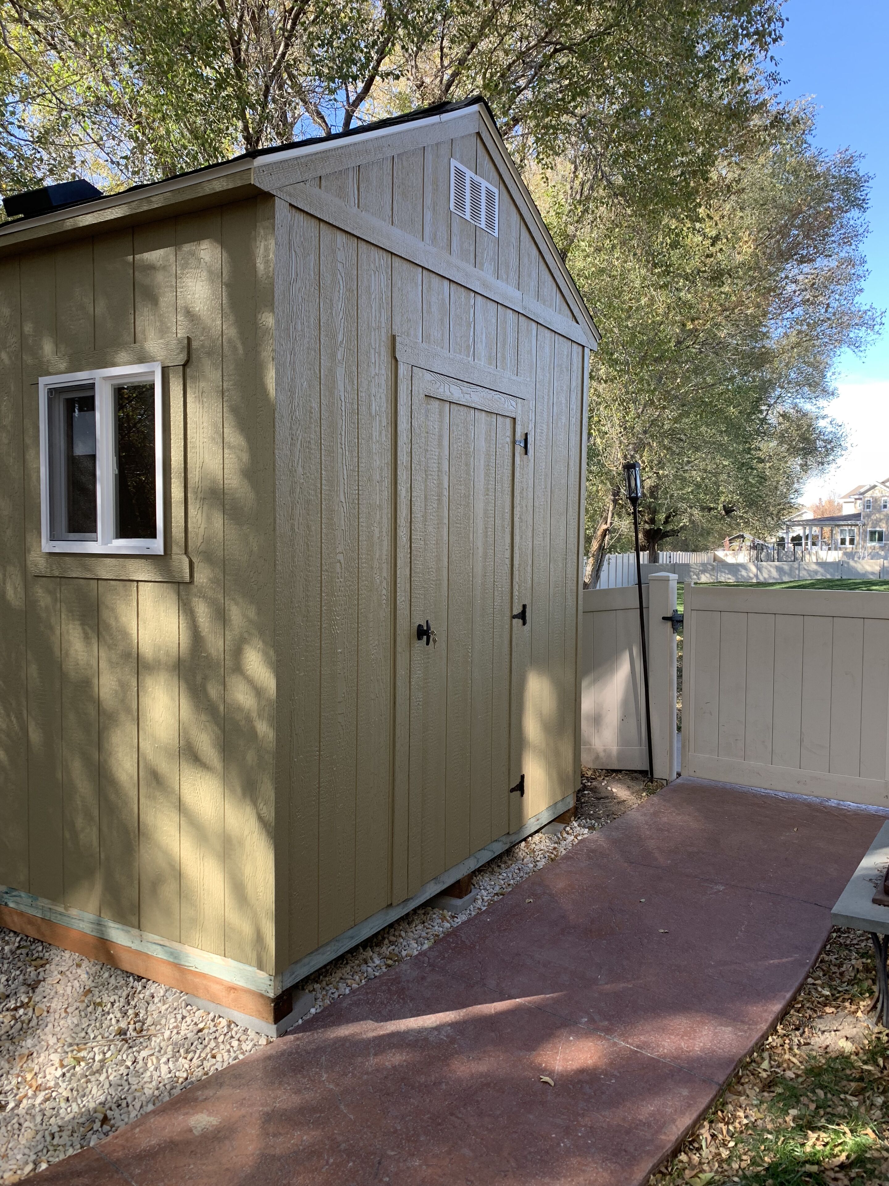 A wooden shed with a window and a door, surrounded by a gravel path and a fence.