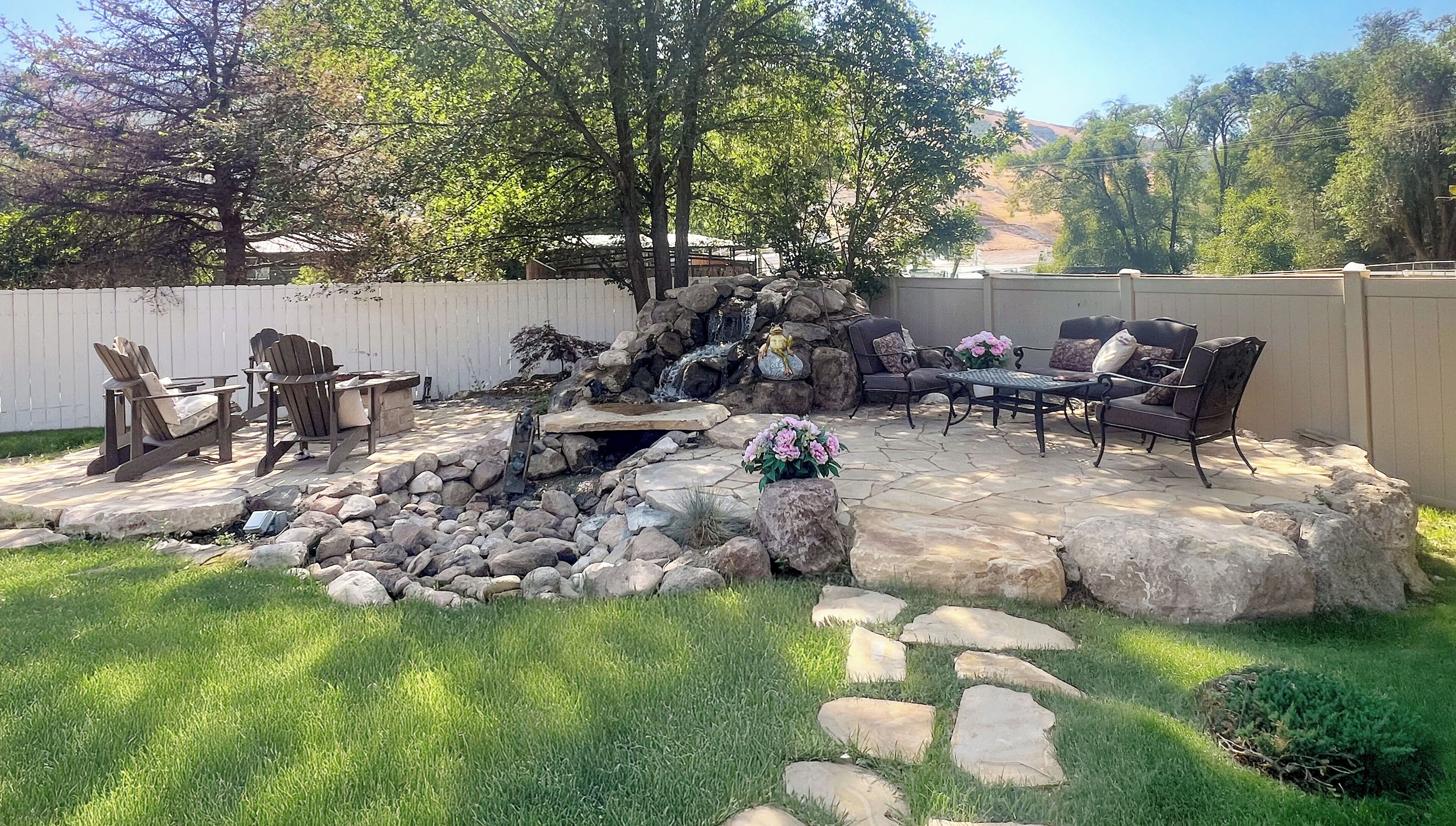 A backyard patio with a stone fire pit, seating area, and a small waterfall feature.
