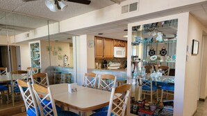 A dining area with a white table and blue cushioned chairs, a kitchen with wooden cabinets, and a glass partition with kitchenware.