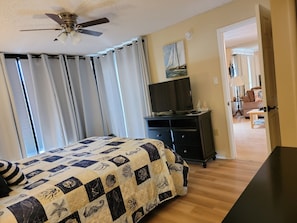 Une chambre à coucher avec un lit, un ventilateur de plafond, un téléviseur posé sur une commode et un tableau accroché au mur.
