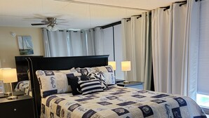 A neatly made bed with a quilt, a bedside table with a lamp, a ceiling fan, and a window with curtains.