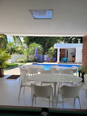 Un área junto a la piscina con una mesa de vidrio y sillas blancas, una piscina con azulejos azules y vistas a una casa moderna con balcón.