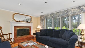 A living room with a fireplace, a sofa, a coffee table, and a large window with a view of greenery. - Chatham-Walk to Ridgevale Beach, Winter Rental Option (Chatham)