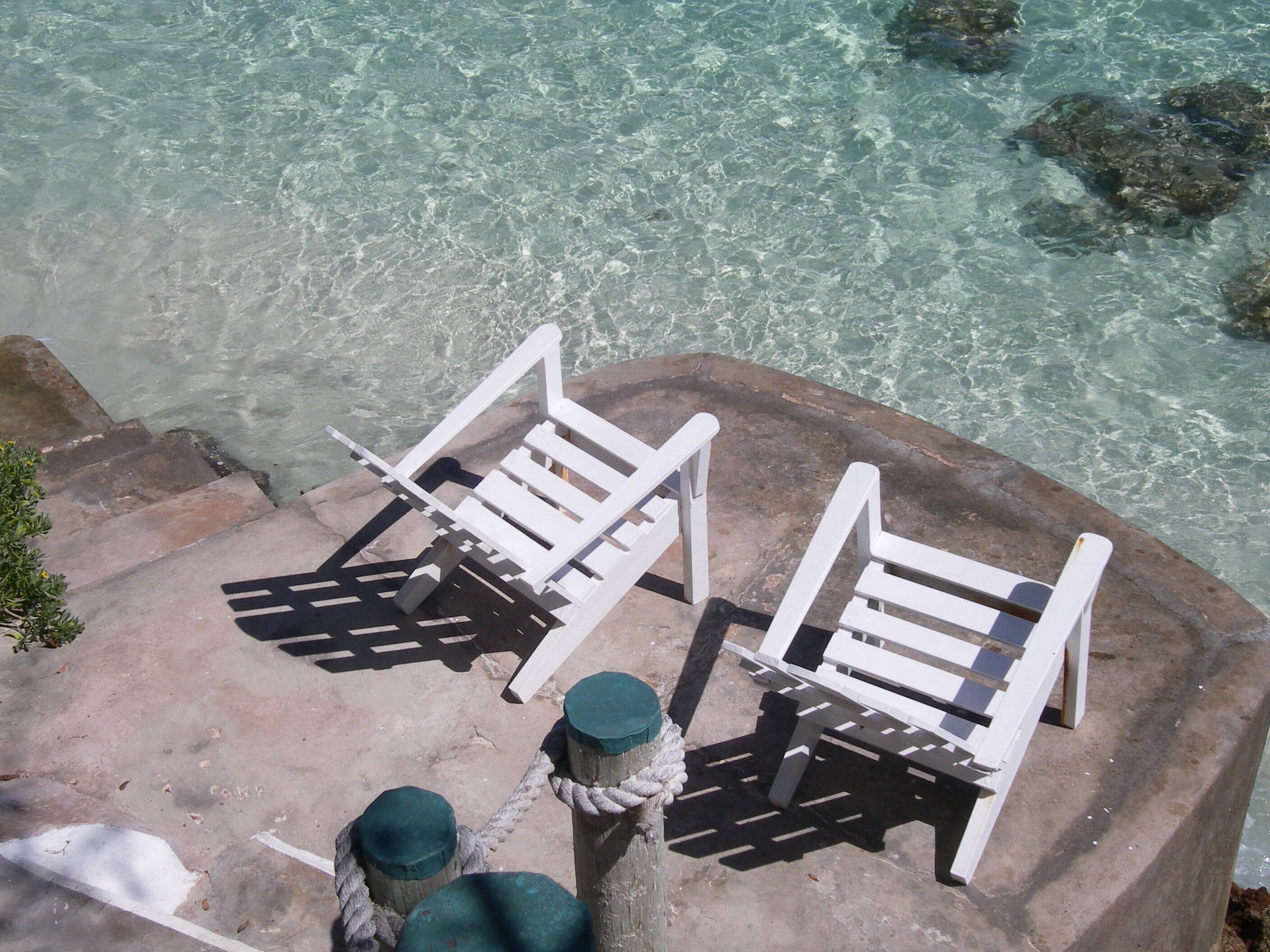 Two white lounge chairs with wooden slats, positioned on a concrete ledge overlooking clear blue water.