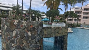 A stone fountain in a pool with palm trees and a building in the background. - Amazing Condo in Cowpet Bay West, Steps From the Beach, Pool and Restaurants (St. Thomas)