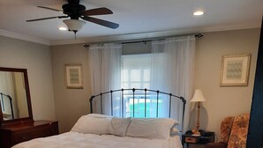Un dormitorio con cama, ventilador de techo, mesita de noche con lámpara, una silla y una ventana con cortinas.