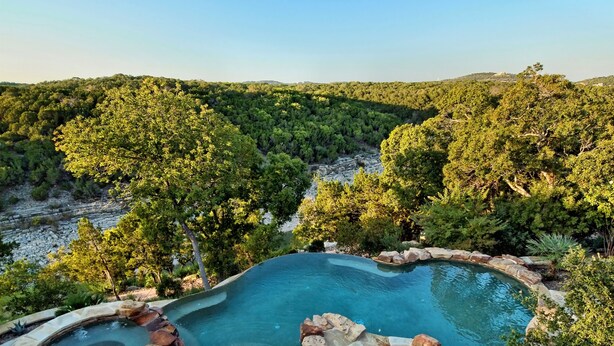 Une piscine au design naturel, avec des pierres apparentes, entourée d’arbres et offrant une vue sur un paysage vallonné.