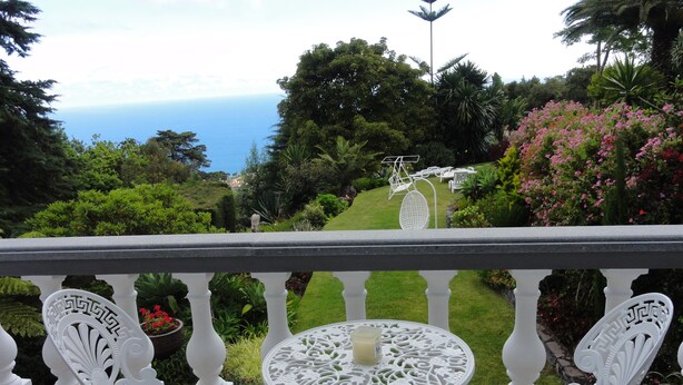 A balcony with white ornate chairs and a table, overlooking a lush garden and the ocean. - Madeiran Quinta with a wonderful 4000 m2 Botanical Garden with private pool (S.Gonçalo)