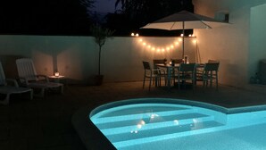 Ein Pool mit gewölbter Kante und ein Tisch mit Stühlen unter Lichterketten.