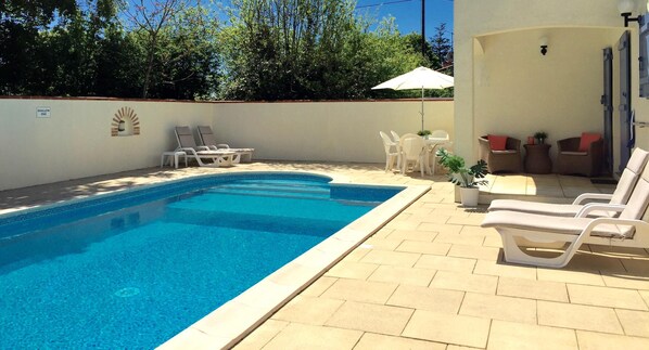 Une piscine entourée d’une terrasse carrelée et d’un petit muret, avec des chaises longues et une table à proximité.