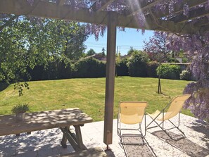 Une terrasse couverte avec une table en bois et deux chaises, donnant sur une cour verdoyante avec des arbres et une maison en arrière-plan.