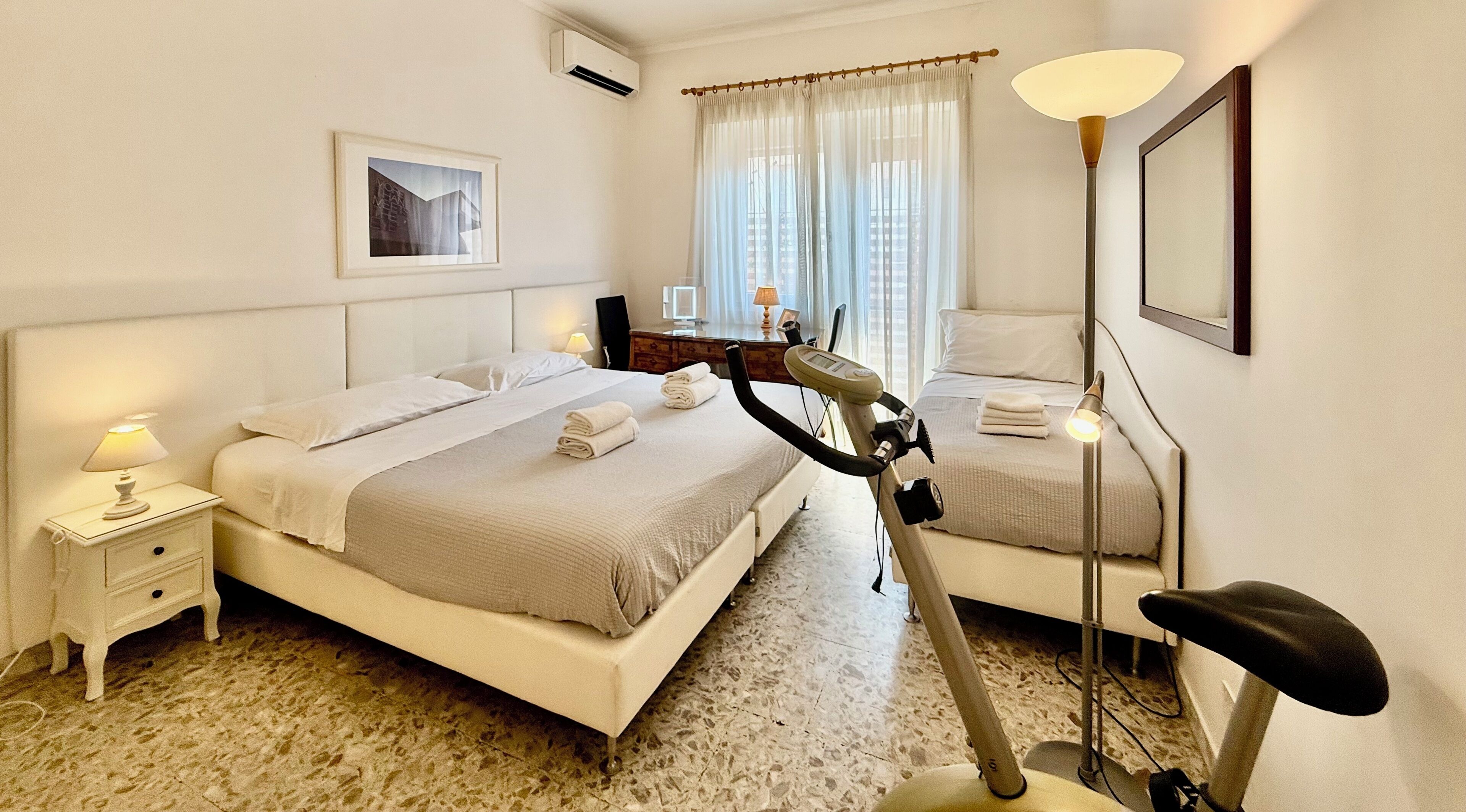 Une chambre d’hôtel avec un lit, un vélo d’exercice, des lampes de chevet, un tableau au mur et une fenêtre avec des rideaux.