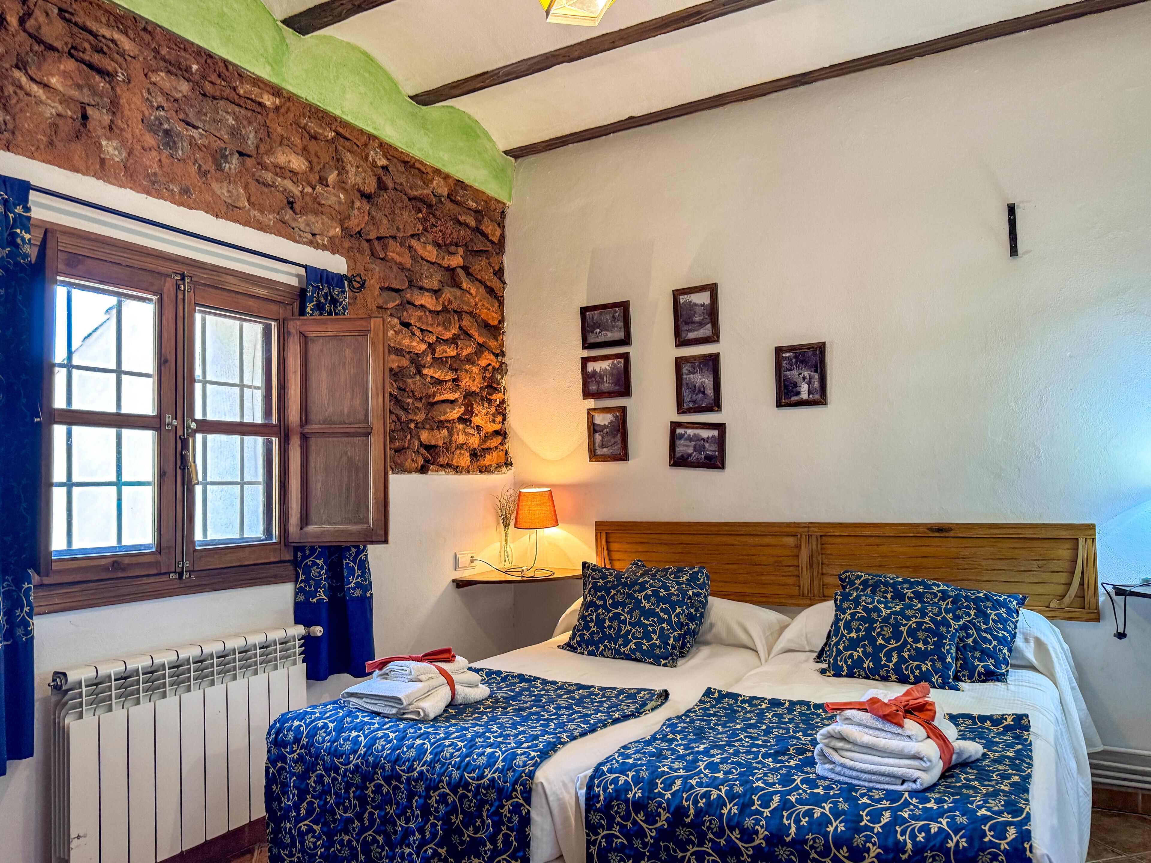 A bedroom with two beds, a stone wall, a wooden ceiling, and a window with curtains.