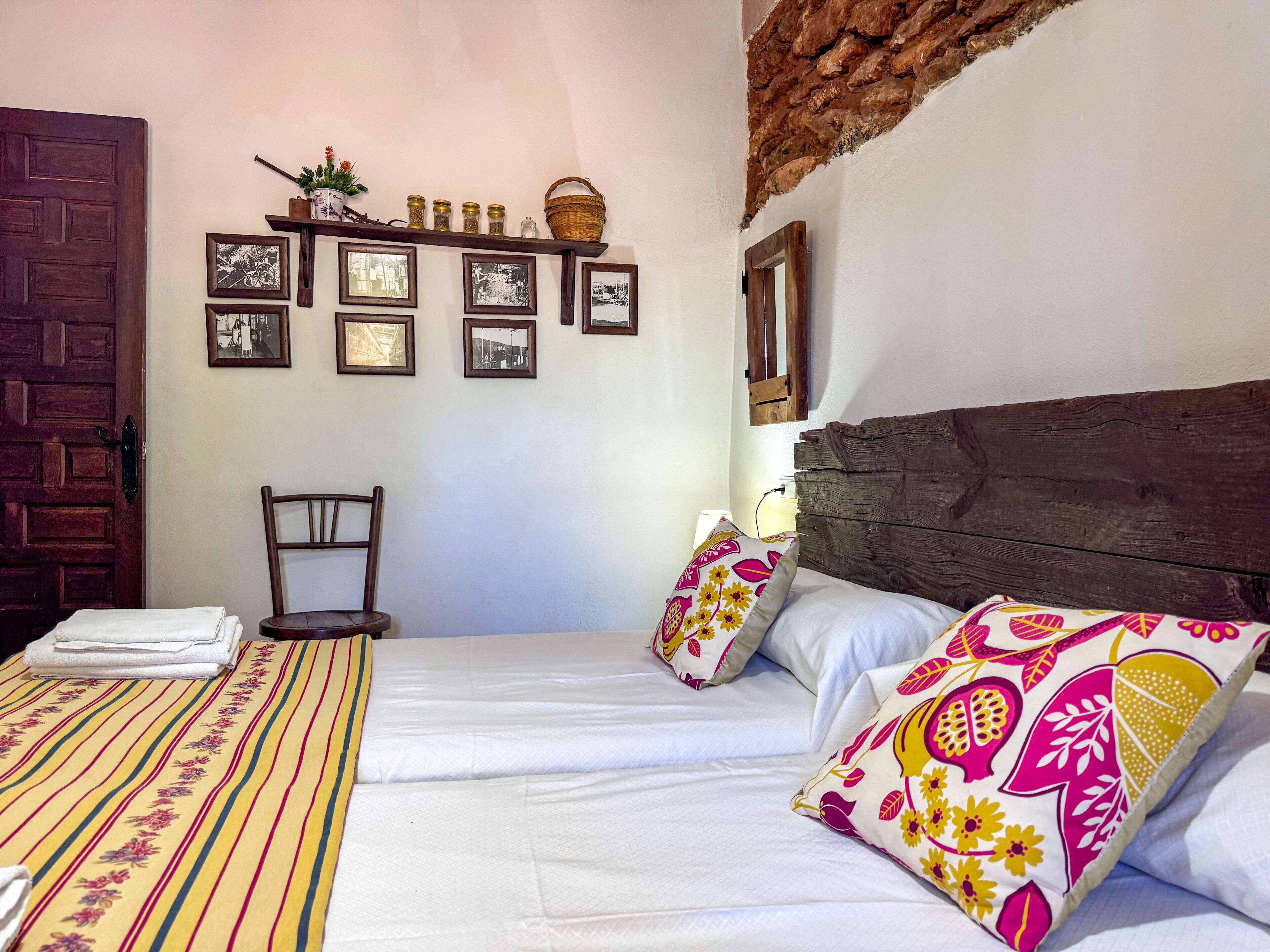 Une chambre avec une tête de lit en bois, un lit recouvert de draps blancs, une couvre-lit à rayures et plusieurs coussins aux motifs floraux. On y trouve également une chaise en bois, une étagère avec des photos encadrées et un panier accroché au mur.