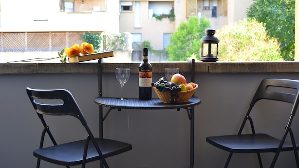 Un balcón con dos sillas, una mesa, una botella de vino, copas y una cesta de fruta.