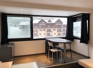 Een kamer met een groot raam, een houten tafel, twee stoelen, een kleine televisie en een bank.