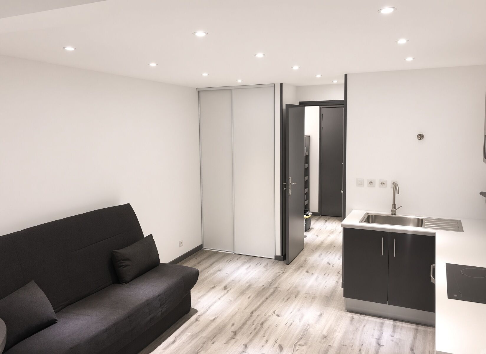A modern living space with a sofa, a kitchen area, and a sliding door leading to another room.
