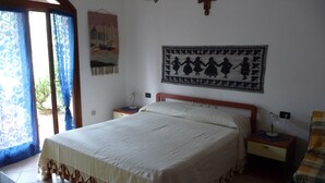 Un dormitorio con una cama grande, una mesita de noche, una mesilla auxiliar, una cruz en la pared y una ventana con cortinas.