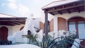 Un edificio blanco con puertas y ventanas de madera, un porche cubierto con sillas y una escalera que sube.