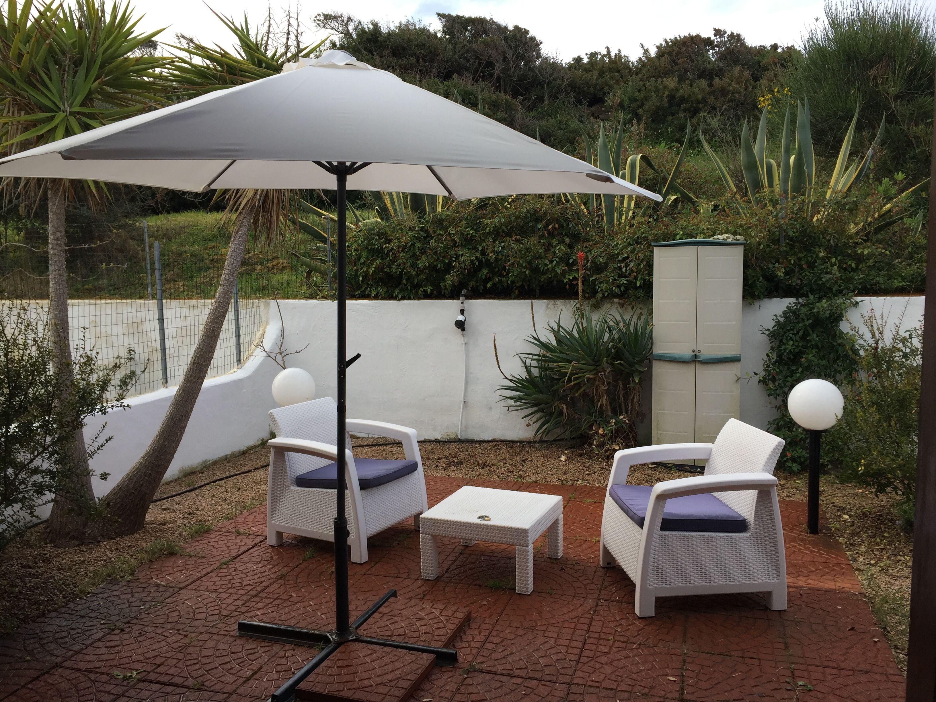Une terrasse extérieure avec deux fauteuils en osier et une table, un parasol blanc et un meuble blanc.