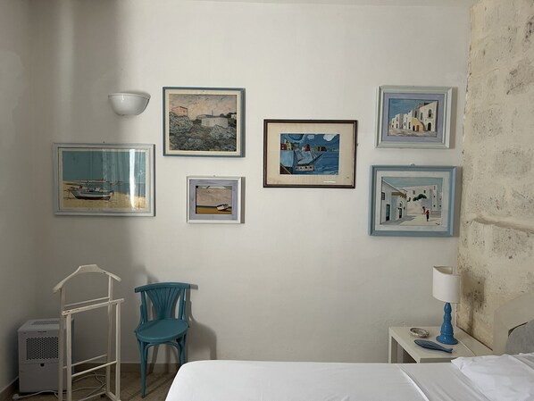 Quarto de hotel com cama, escrivaninha com luminária, cadeira e quatro obras de arte emolduradas na parede.