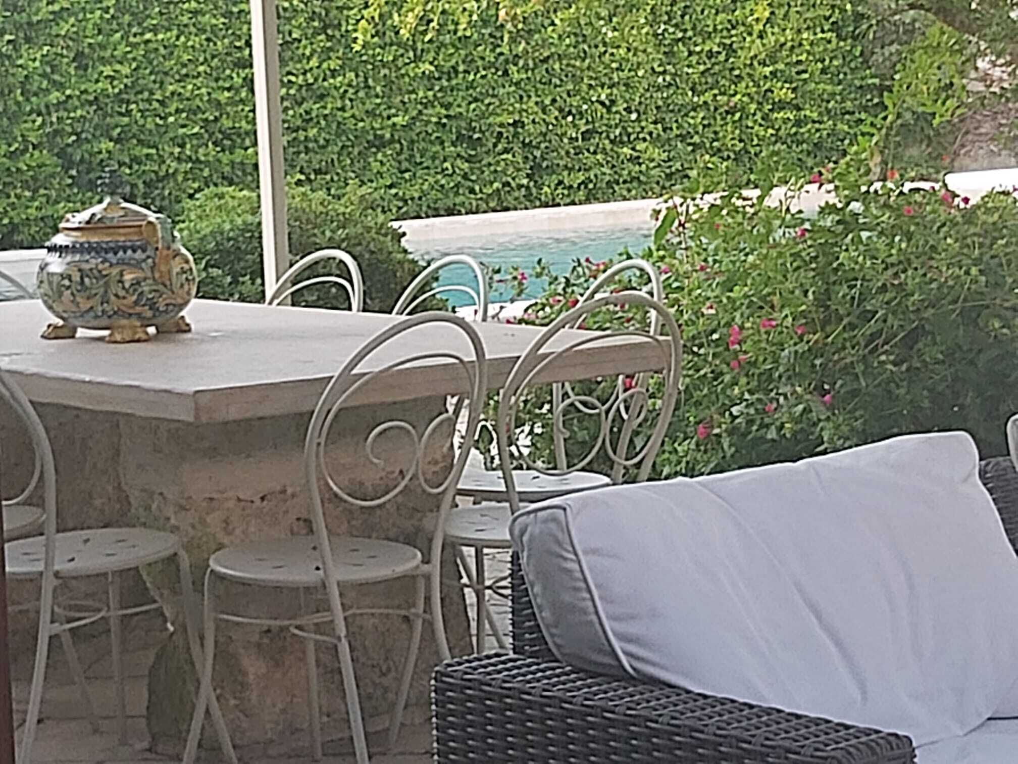 An outdoor seating area with a stone table and metal chairs, surrounded by greenery and a pool.