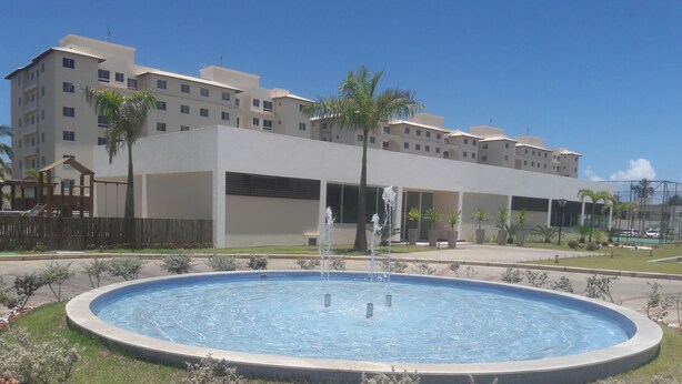 Una piscina con una fuente, rodeada por un jardín ajardinado con rocas y árboles pequeños, frente a un edificio de varios pisos con balcones.