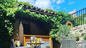 Ein Sitzbereich im Freien mit Korbstühlen und einem Tisch, eine Holzpergola mit Kletterpflanzen und eine Steintreppe im Hintergrund.