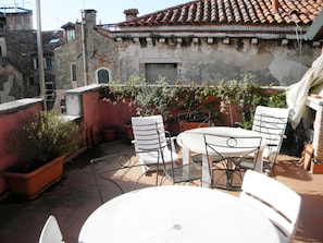 Un piccolo patio esterno con un tavolo rotondo e quattro sedie, circondato da piante in vaso e un muro in mattoni rossi.