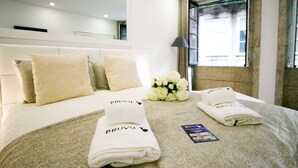 Una cama bien hecha con una colcha blanca, dos toallas dobladas y un ramo de flores blancas.