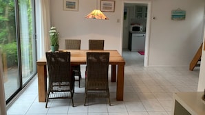 Un comedor con mesa de madera y sillas de mimbre, una lámpara colgante y vistas a la cocina a través de una puerta abierta.
