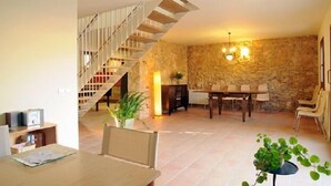 Amplia sala de estar con pared de piedra, escalera de madera, mesa de comedor con sillas y chimenea.