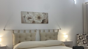 En pænt redt seng med et beige dynebetræk, to puder og en gavl med en lampe. Et billede af blomster hænger på væggen over sengen, og et natbord med en lampe er placeret til venstre.