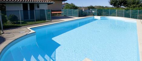 A rectangular swimming pool with a clear blue water surface, surrounded by a beige tiled deck and a green fence.