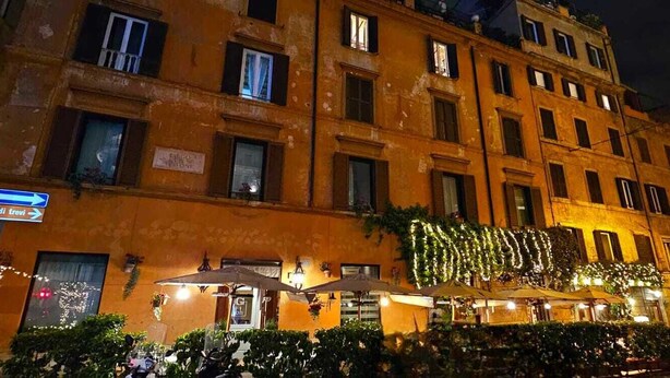 Un edificio histórico con múltiples ventanas y balcones, mobiliario exterior con sombrillas y un letrero que indica una fuente cercana.