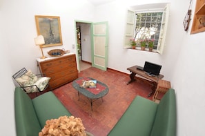 Ein gemütliches Zimmer mit einem grünen Sofa, einer Holzkommode, einem Korbtisch mit einem Buch, einem Fernseher auf einem Holzschrank und einem Fenster mit Topfpflanzen.