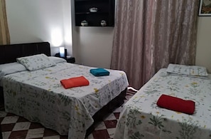 Ein Zimmer mit zwei Betten, jeweils mit floraler Bettwäsche und einem roten und blauen Handtuch. Ein Nachttisch mit Lampe, ein Regal mit Dekorationen und ein Fenster mit Vorhängen.