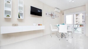 A modern living room with a flat-screen TV, white shelves with decorative items, a dining area with a glass table and white chairs, and a large window offering a view of the outside. - Rent Corro X32 (Córdoba)